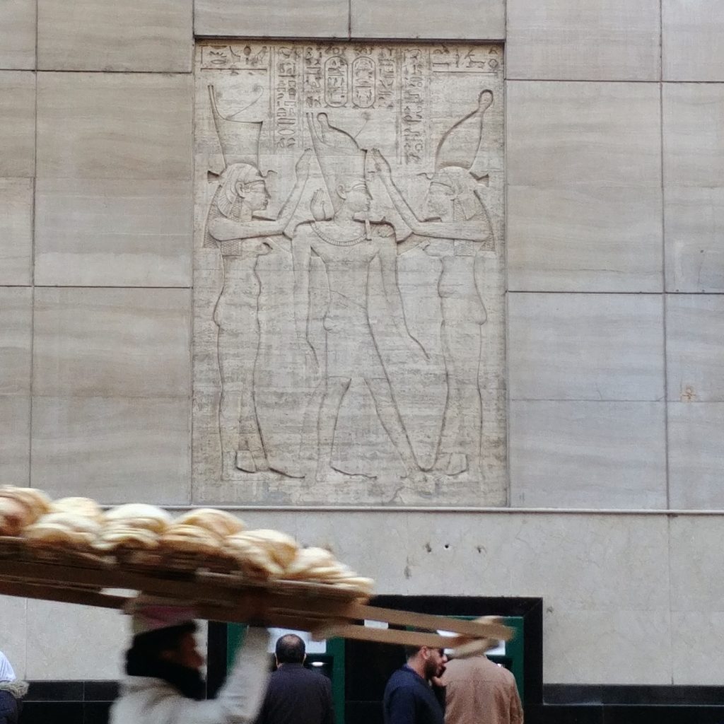 an egyptian man carries fresh bread in the street, there is wall decoration in the background with Egyptian hieroglyphs