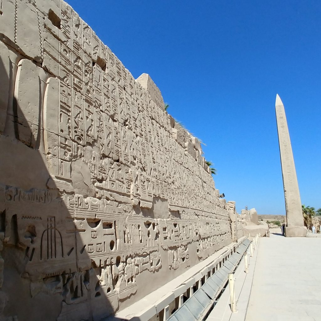 wall filled with hieroglyphs and a massive obelisk in the background at Karnak Temple - Luxor