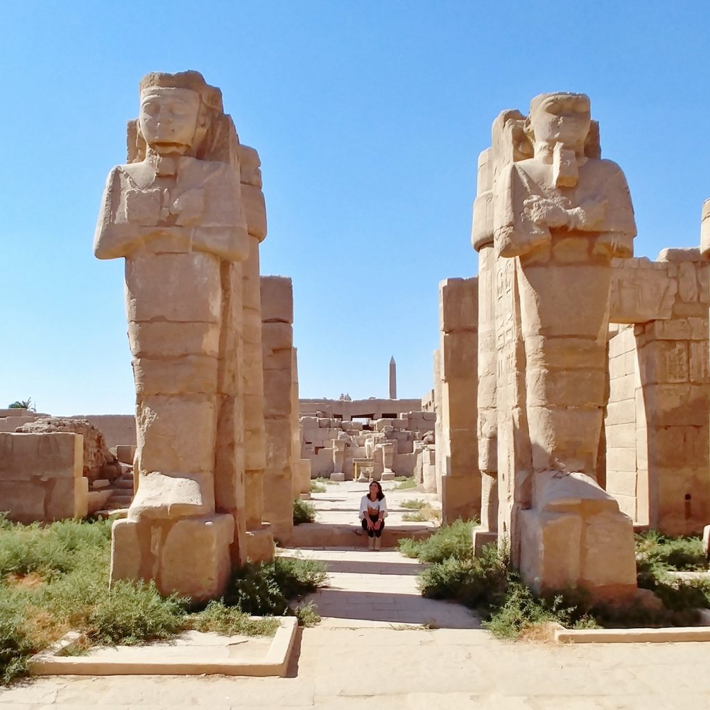 a girl is sittign next to 2 giant statues at Karnak Temple - Luxor